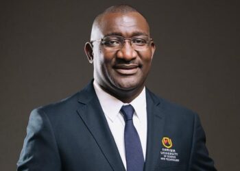 Frednard Gideon - The Brief | Namibia's Leading Business & Financial News Professional head-and-shoulders portrait of a smiling man in a navy suit and tie, wearing glasses, with a Namibia University of Science and Technology logo on his blazer.