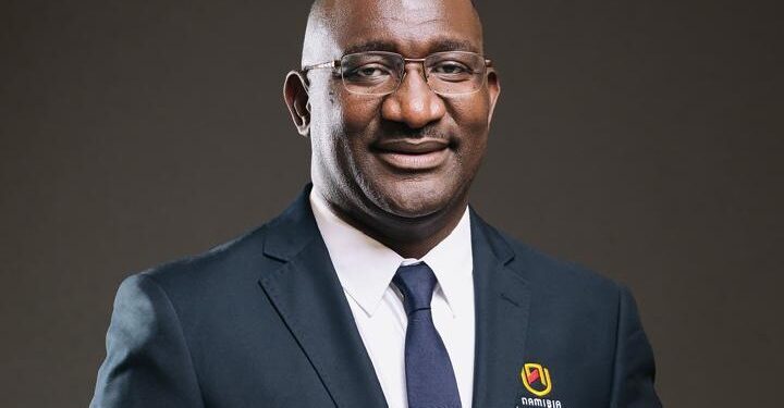 Professional head-and-shoulders portrait of a smiling man in a navy suit and tie, wearing glasses, with a Namibia University of Science and Technology logo on his blazer.