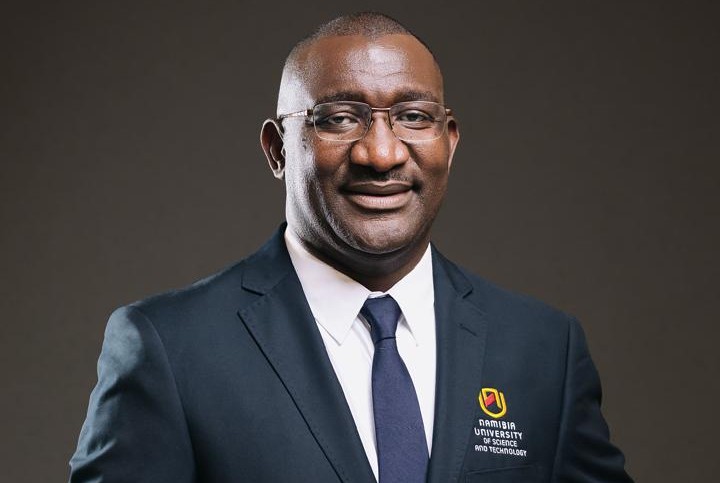 Professional head-and-shoulders portrait of a smiling man in a navy suit and tie, wearing glasses, with a Namibia University of Science and Technology logo on his blazer.