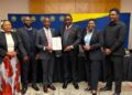 Six professionals in business attire posing for a photo at a ceremony, with a man in the center holding a certificate.
