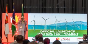 Speaker at a podium giving a speech at an official launch event, with flags and a banner that reads 'Official Launch of the Real Test Project' behind her.