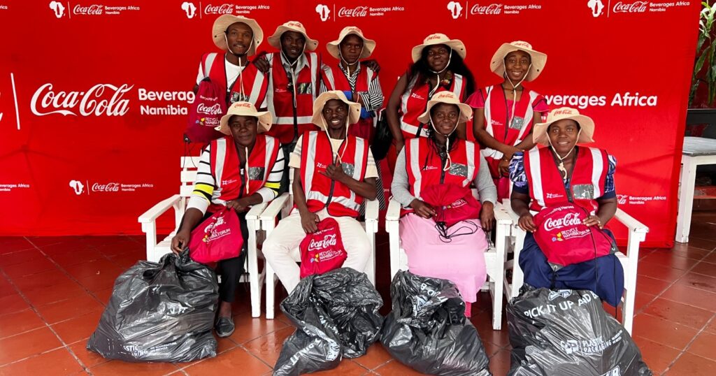 Group of Coca-Cola Namibia volunteers in red vests and hats pose for photo with trash bags at a Beverages Africa event backdrop