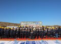 Group photo of dozens of workers in navy and tan uniforms wearing hard hats on a red carpet at a mining site, with a banner in the background reading Sinomine Tsumeb Multi-Metal Recovery.