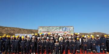 Group photo of dozens of workers in navy and tan uniforms wearing hard hats on a red carpet at a mining site, with a banner in the background reading Sinomine Tsumeb Multi-Metal Recovery.