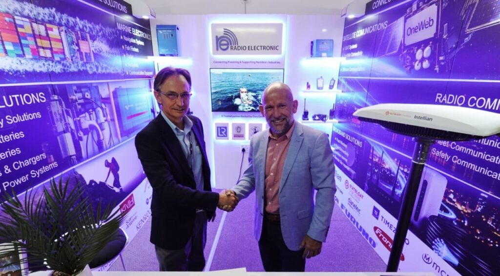 Two men in business attire shake hands at a tech expo booth with blue-lit banners and branding.