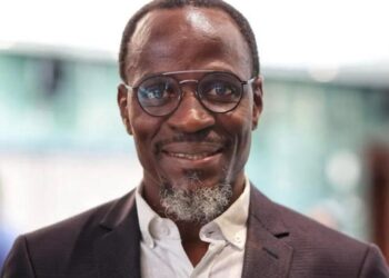 Fillemon Wise - The Brief | Namibia's Leading Business & Financial News Professional man with glasses in a dark blazer and white shirt, smiling at the camera in a bright indoor setting.