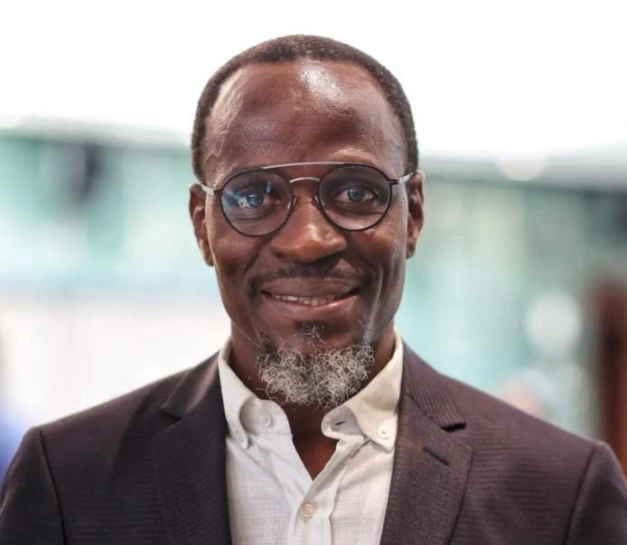 Professional man with glasses in a dark blazer and white shirt, smiling at the camera in a bright indoor setting.