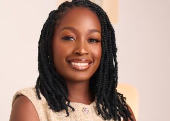 Close-up portrait of a smiling Black woman with long black braids and a beige textured top against a light background.