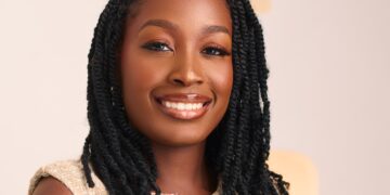 Close-up portrait of a smiling Black woman with long black braids and a beige textured top against a light background.