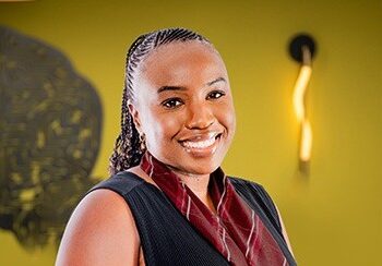 Helvi Fillipus - The Brief | Namibia's Leading Business & Financial News Smiling woman in a dark vest and maroon scarf stands against a yellow wall.