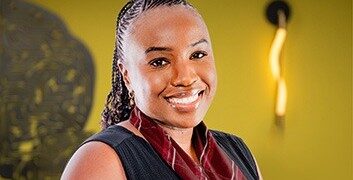 Smiling woman in a dark vest and maroon scarf stands against a yellow wall.