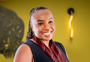 Smiling woman in a dark vest and maroon scarf stands against a yellow wall.