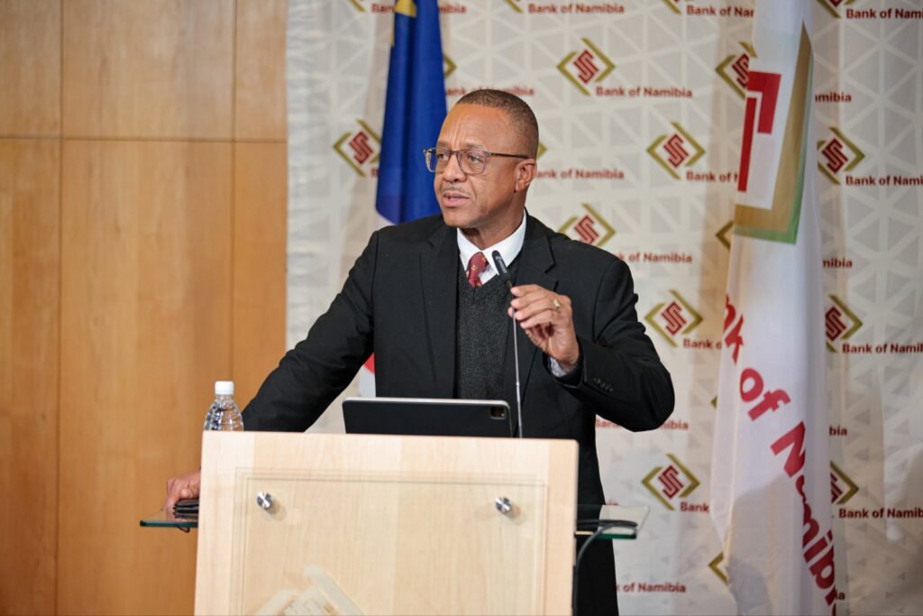 Uanguta 20 - The Brief | Namibia's Leading Business & Financial News Man in a dark suit and glasses gives a speech at a wooden podium with a microphone and tablet in front, Bank of Namibia backdrop behind him.