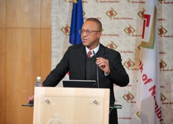 Man in a dark suit and glasses gives a speech at a wooden podium with a microphone and tablet in front, Bank of Namibia backdrop behind him.