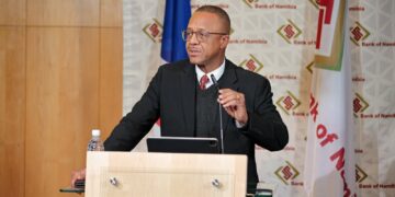 Man in a dark suit and glasses gives a speech at a wooden podium with a microphone and tablet in front, Bank of Namibia backdrop behind him.
