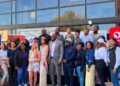Diverse group of people posing outside a glass-front building for a community event, several wearing sashes and smiling at the camera.