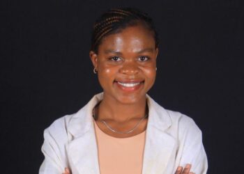 Apolonia H Ndakolo - The Brief | Namibia's Leading Business & Financial News Smiling woman in a light blazer over a peach top, posing with arms crossed against a dark background.
