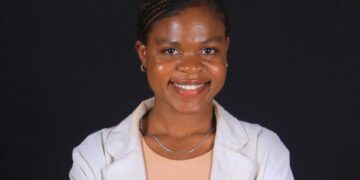 Smiling woman in a light blazer over a peach top, posing with arms crossed against a dark background.