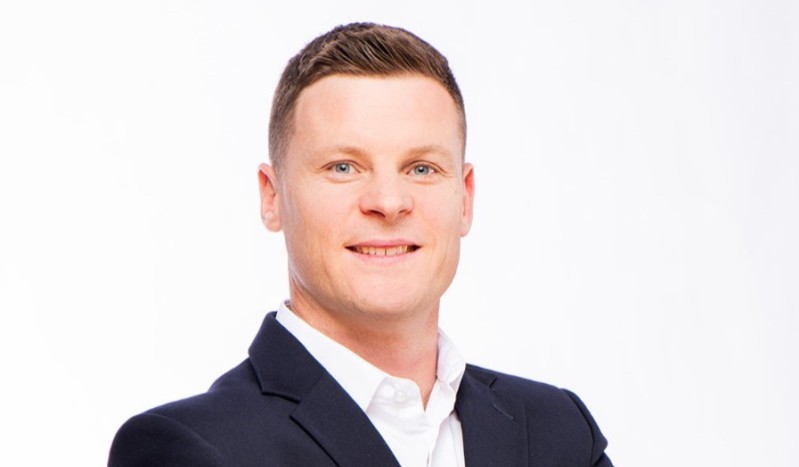 Professional headshot of a smiling man in a dark suit and white shirt against a light background.