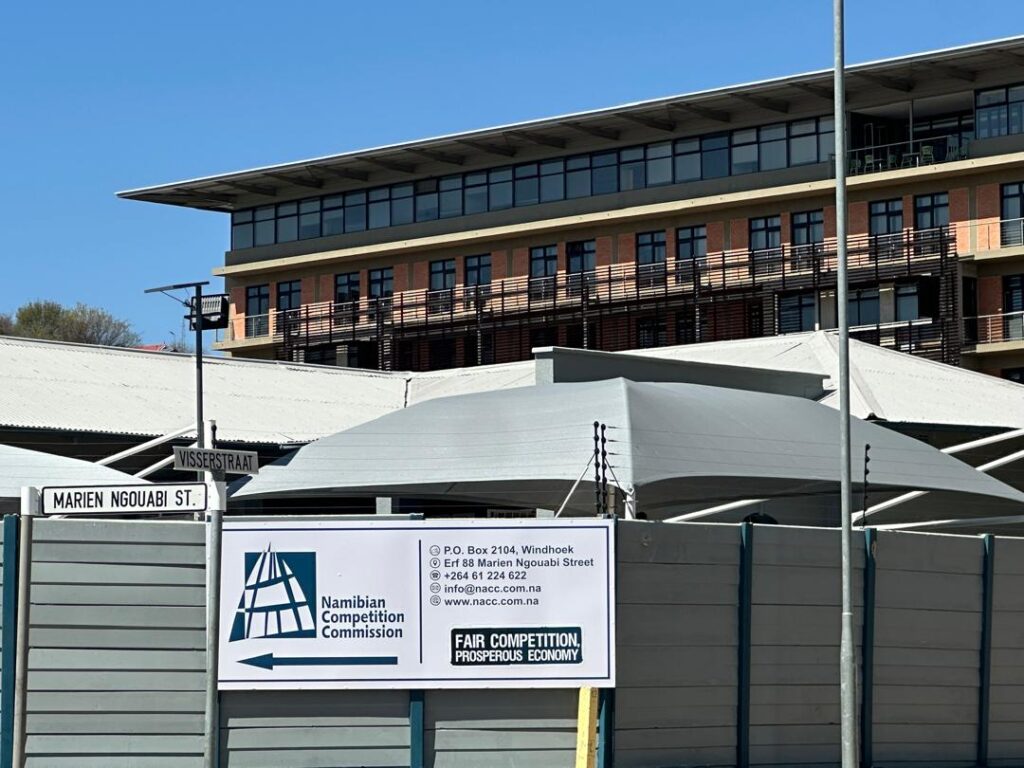Namibia Competition Commission - The Brief | Namibia's Leading Business & Financial News Construction-site fence with a Namibian Competition Commission banner in front of a modern brick building; street signs show Marien Ngouabi St and Wisserstraat.