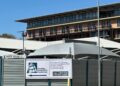 Namibia Competition Commission - The Brief | Namibia's Leading Business & Financial News Construction-site fence with a Namibian Competition Commission banner in front of a modern brick building; street signs show Marien Ngouabi St and Wisserstraat.