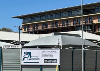 Namibia Competition Commission - The Brief | Namibia's Leading Business & Financial News Construction-site fence with a Namibian Competition Commission banner in front of a modern brick building; street signs show Marien Ngouabi St and Wisserstraat.