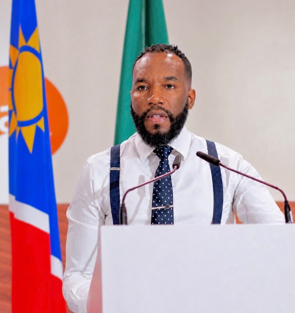 Man wearing a white shirt, dark polka-dot tie and suspenders speaks at a podium, with national flags behind him.