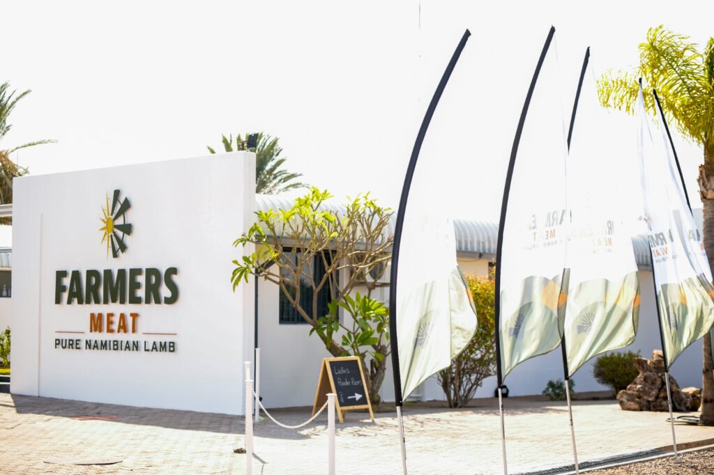 White storefront wall with FARMERS MEAT sign and Pure Namibian Lamb tagline, palm trees behind, plus tall fabric flags along the walkway nearby.