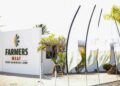 HARTLIEF - The Brief | Namibia's Leading Business & Financial News White storefront wall with FARMERS MEAT sign and Pure Namibian Lamb tagline, palm trees behind, plus tall fabric flags along the walkway nearby.