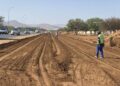 WhatsApp Image 2026-08-17 at 71301 PM - The Brief | Namibia's Leading Business & Financial News Construction workers in high-visibility vests along a dirt road under construction beside a busy highway; muddy tire tracks, piles of soil, and distant hills.