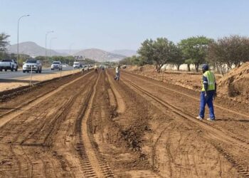 WhatsApp Image 2026-08-17 at 71301 PM - The Brief | Namibia's Leading Business & Financial News Construction workers in high-visibility vests along a dirt road under construction beside a busy highway; muddy tire tracks, piles of soil, and distant hills.