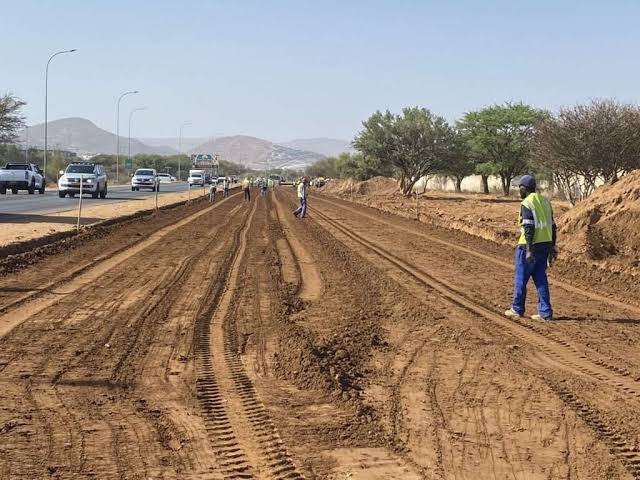 WhatsApp Image 2026-08-17 at 71301 PM - The Brief | Namibia's Leading Business & Financial News Construction workers in high-visibility vests along a dirt road under construction beside a busy highway; muddy tire tracks, piles of soil, and distant hills.