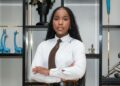 Ngurije Tjiroze - The Brief | Namibia's Leading Business & Financial News Professional woman with arms crossed, wearing a white shirt and brown tie, standing in front of a display shelf with blue giraffe sculptures behind her.