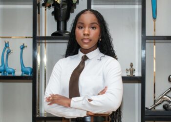 Ngurije Tjiroze - The Brief | Namibia's Leading Business & Financial News Professional woman with arms crossed, wearing a white shirt and brown tie, standing in front of a display shelf with blue giraffe sculptures behind her.