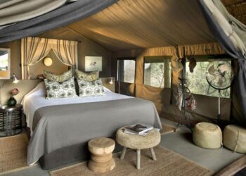 Luxurious safari-glamping bedroom in a canvas tent with a king bed, canopy, and decorative pillows. In-room seating and warm lighting create a cozy retreat at the camp.
