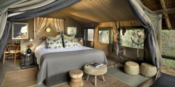 Luxurious safari-glamping bedroom in a canvas tent with a king bed, canopy, and decorative pillows. In-room seating and warm lighting create a cozy retreat at the camp.