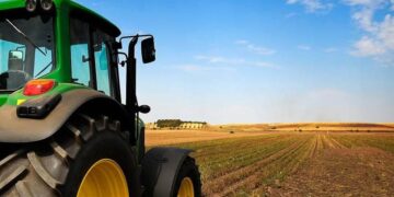 Green tractor with large yellow wheels on a flat farm field under a bright blue sky