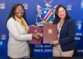 Two women in business attire shake hands while holding signed documents in front of a blue banner with a crest and motto.