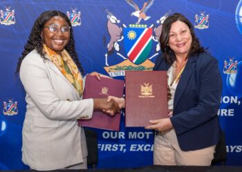 Two women in business attire shake hands while holding signed documents in front of a blue banner with a crest and motto.