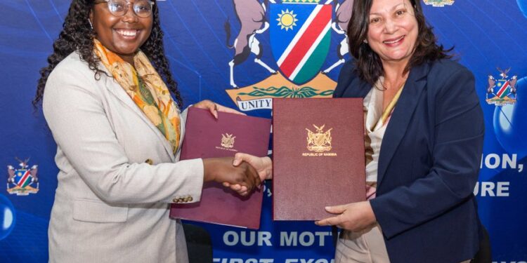 Two women in business attire shake hands while holding signed documents in front of a blue banner with a crest and motto.