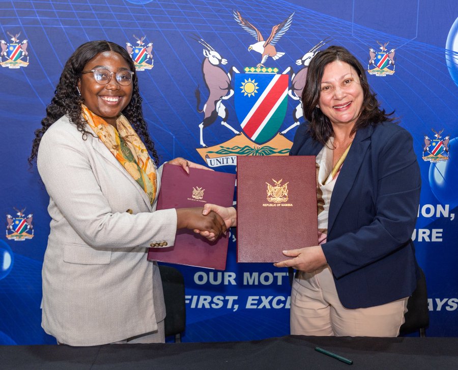 Two women in business attire shake hands while holding signed documents in front of a blue banner with a crest and motto.