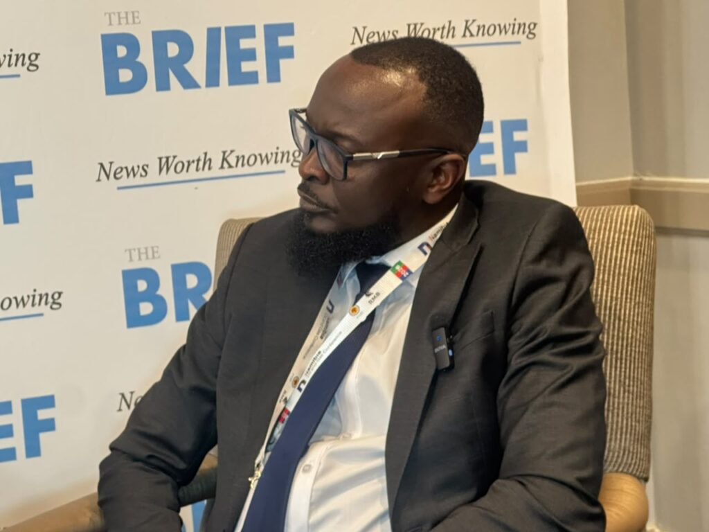 JASON KASUTO3 - The Brief | Namibia's Leading Business & Financial News Side profile of a bearded man in a dark suit and glasses seated at a conference, backdrop reads 'The BRIEF' and 'News Worth Knowing'.