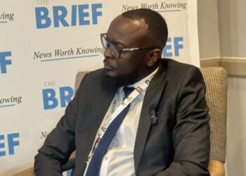 Side profile of a bearded man in a dark suit and glasses seated at a conference, backdrop reads 'The BRIEF' and 'News Worth Knowing'.