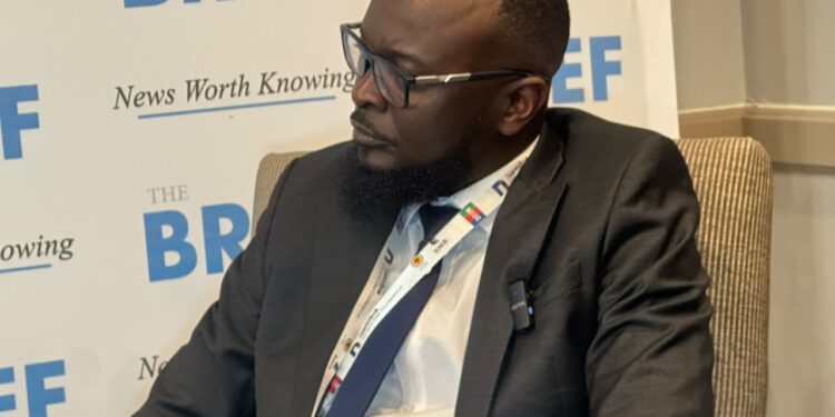 Side profile of a bearded man in a dark suit and glasses seated at a conference, backdrop reads 'The BRIEF' and 'News Worth Knowing'.