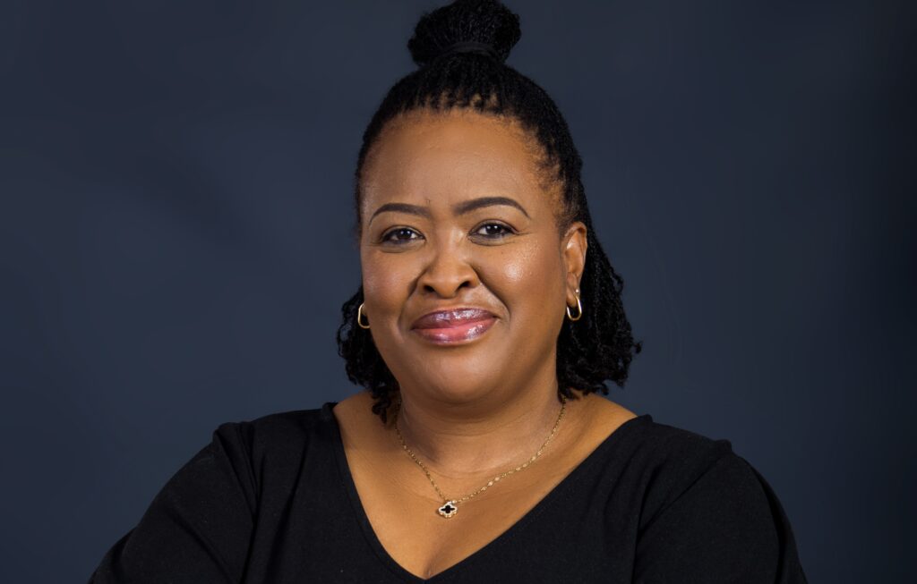 Smiling woman with a black top and gold jewelry against a dark blue studio backdrop.