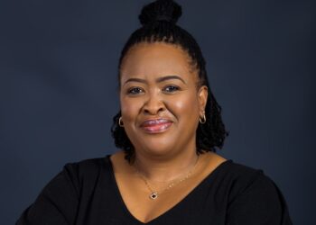 corny zaaruka - The Brief | Namibia's Leading Business & Financial News Smiling woman with a black top and gold jewelry against a dark blue studio backdrop.