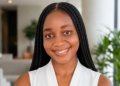 Smiling Black woman with long box braids in a sleeveless white top, indoors with plants in the background.