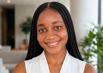 Smiling Black woman with long box braids in a sleeveless white top, indoors with plants in the background.
