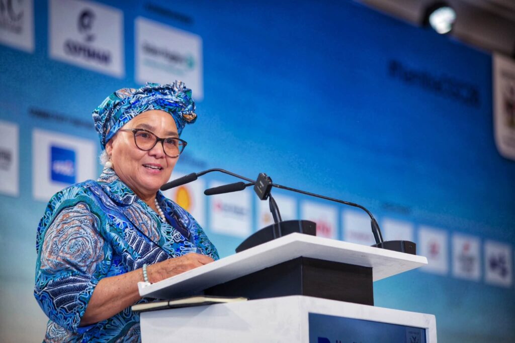 Senior woman in a blue patterned outfit and head wrap speaks at a podium during a conference.