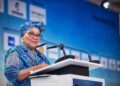 Senior woman in a blue patterned outfit and head wrap speaks at a podium during a conference.